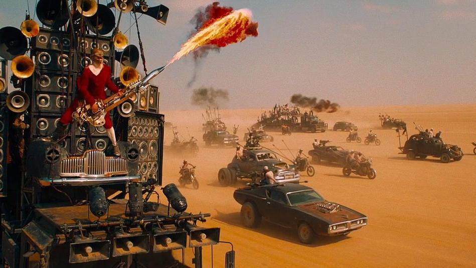 A massive vehicle outfitted with speakers and horns barrels through a desert as a performer in red plays a flame-throwing guitar strapped to its front. Behind it, a convoy of weaponized cars and motorcycles kicks up dust under a blazing sky, evoking a cha