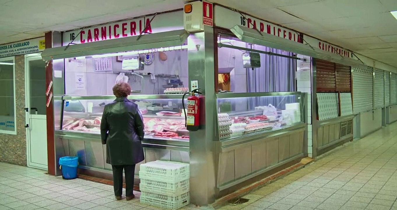 A person in a dark coat stands at the counter of a butcher shop labeled “Carnicería.” Various meats and sausages are displayed, with stacks of eggs to the right. The shop is numbered 16 and features tiled floors, fluorescent lighting, and a fire extinguis