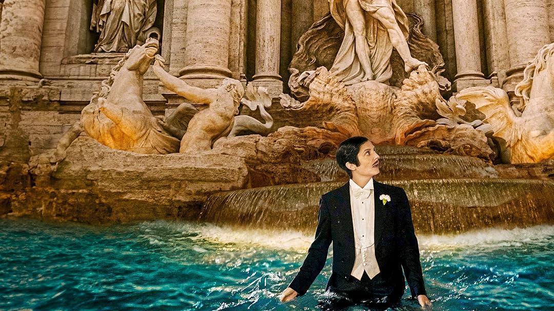 A person in a black tuxedo with a boutonnière stands waist-deep in Rome’s Trevi Fountain, gazing upward. The illuminated Baroque sculptures and cascading water behind them create a dramatic contrast with the formal attire.