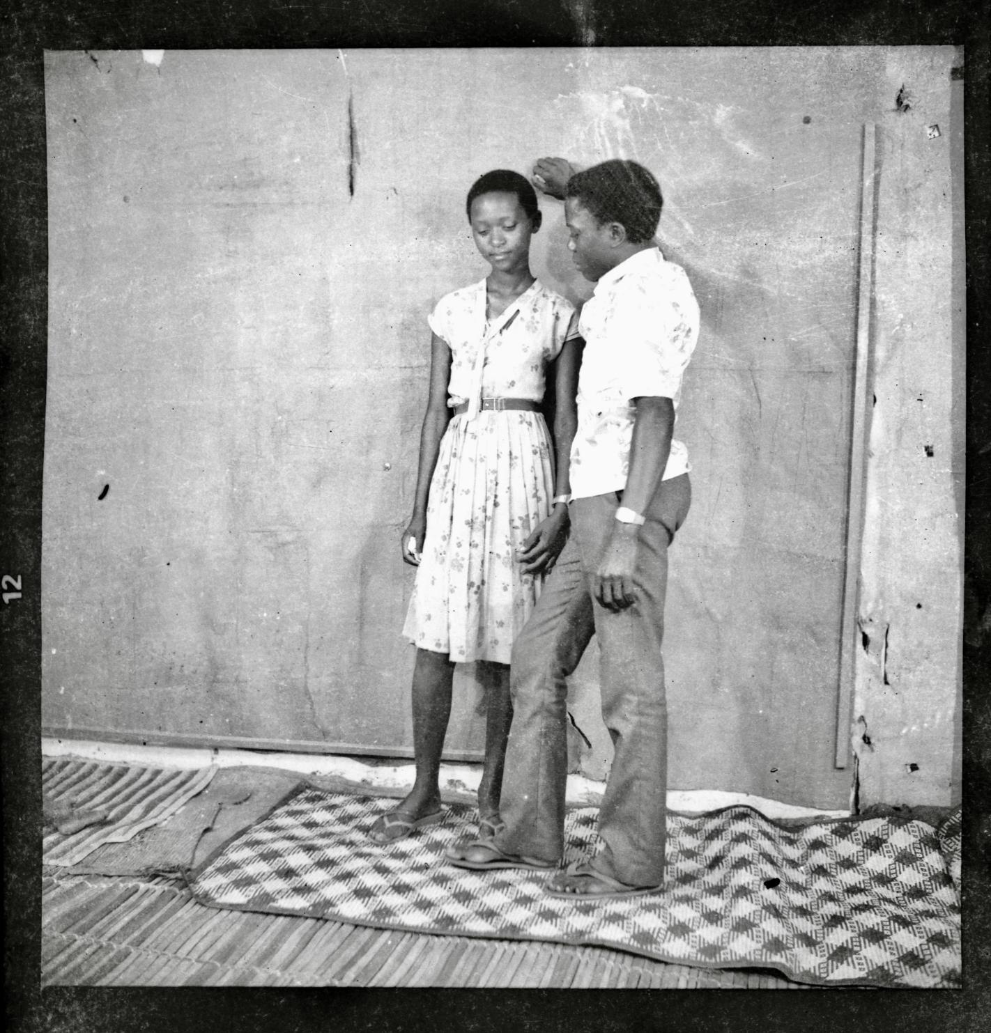 A black-and-white photograph shows two people indoors against a textured backdrop. One stands barefoot on a woven mat wearing a patterned dress, while the other leans slightly toward them with one arm raised, dressed in a short-sleeved shirt and trousers.