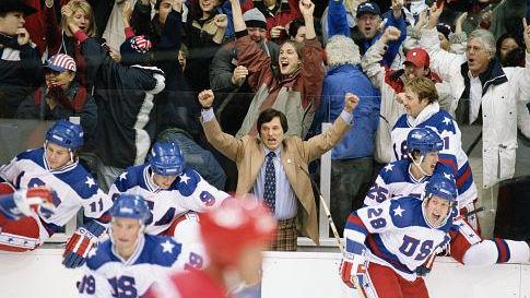 Players in white USA hockey jerseys cheer from the bench as fans erupt behind them. A man in a tan jacket and tie raises his fists in celebration. The scene captures a moment of triumph and national pride during an intense game.