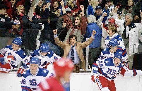 Players in white USA hockey jerseys cheer from the bench as fans erupt behind them. A man in a tan jacket and tie raises his fists in celebration. The scene captures a moment of triumph and national pride during an intense game.
