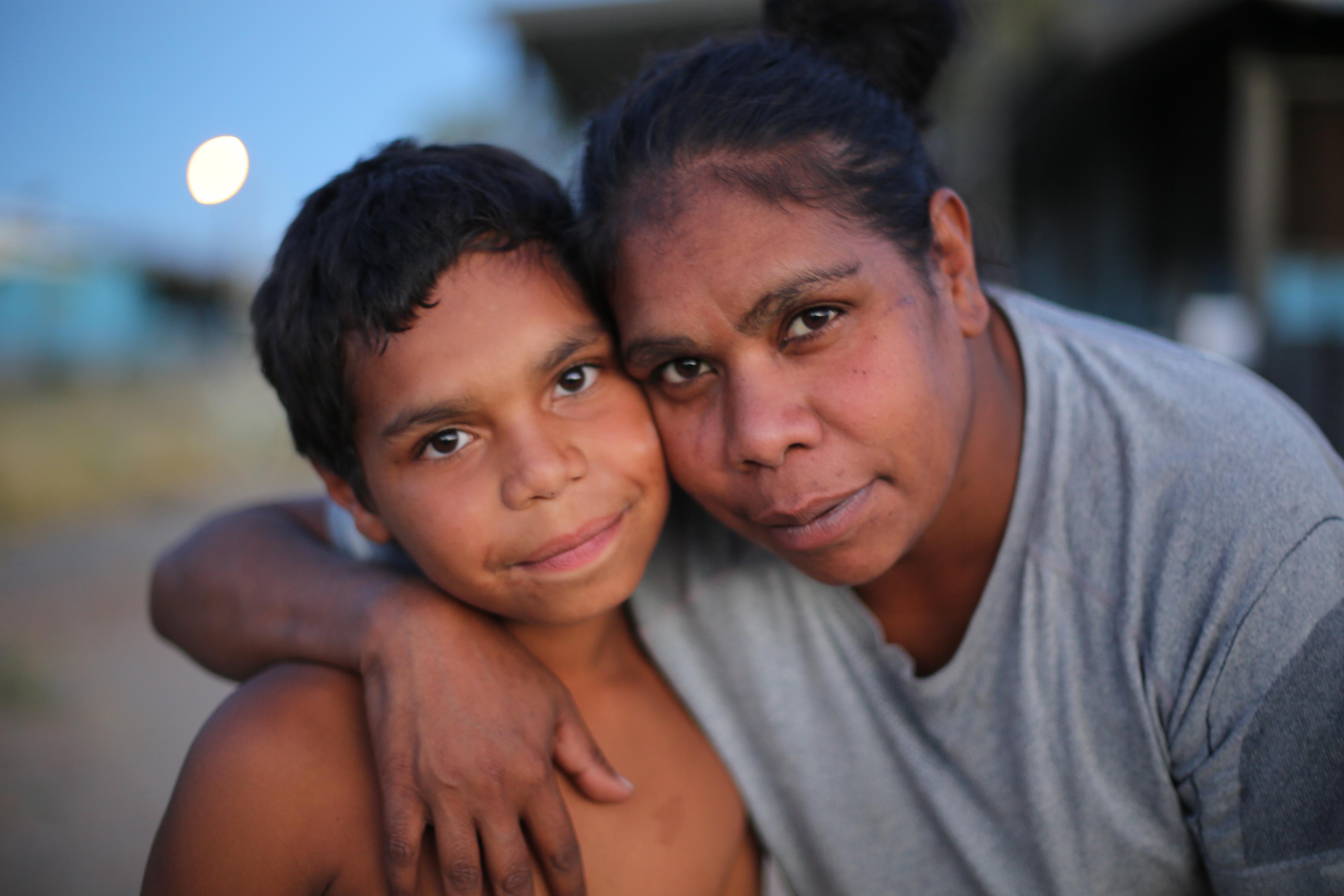 Two people embrace closely with their faces touching, looking directly at the camera. The softly blurred background suggests an outdoor setting during twilight, highlighting a moment of intimacy and connection.