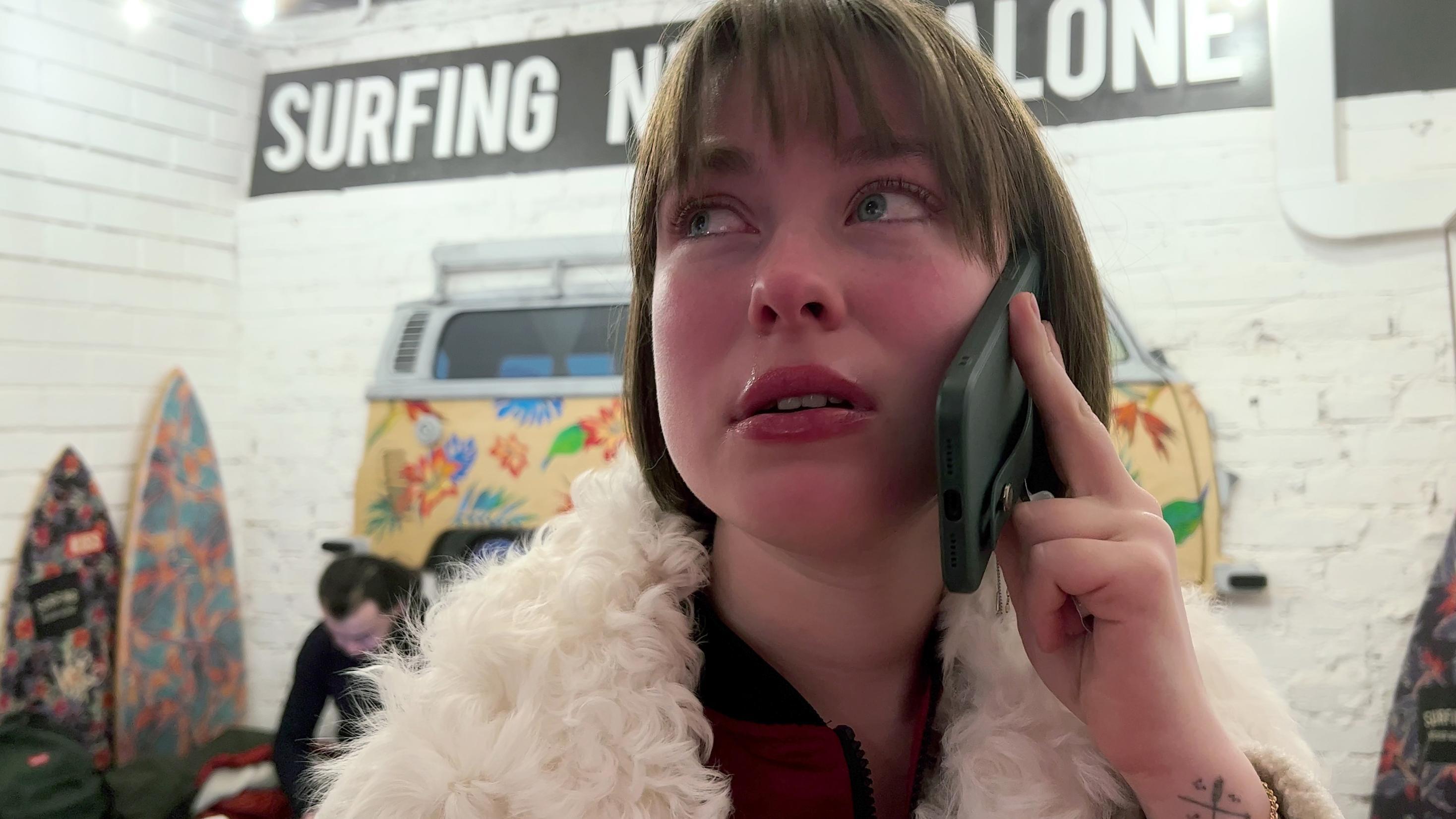 A person in a fluffy white coat holds a smartphone to their ear, appearing serious. Behind them, surfboards lean against a white brick wall and a colorful mural of a van with tropical flowers reads “SURFING NOT ALONE.” 