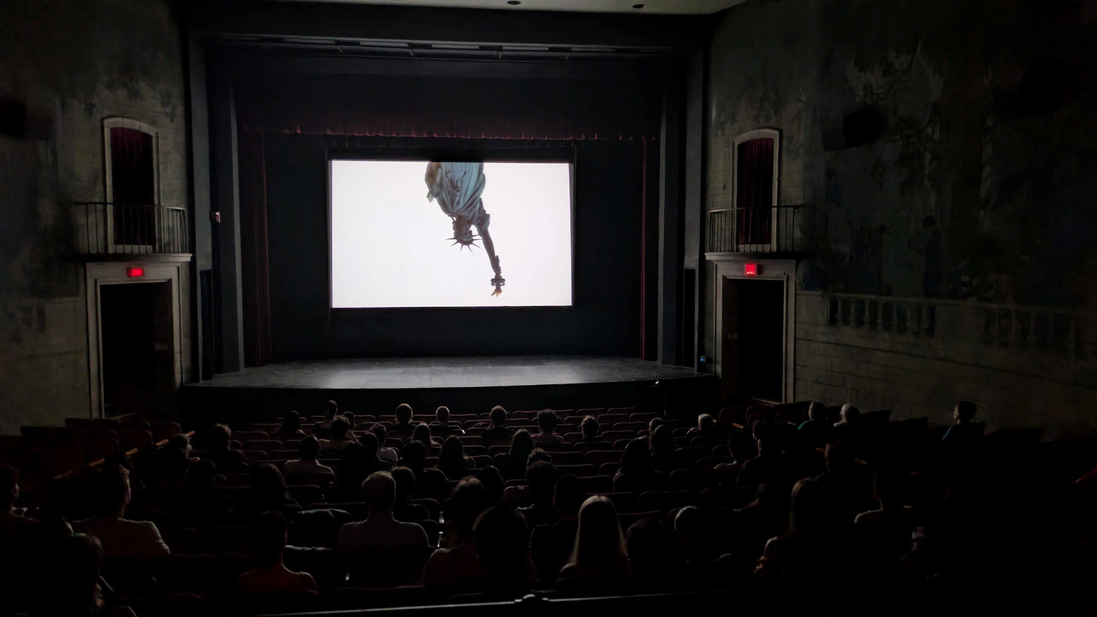 Inside a darkened movie theater, audience members watch a film showing an upside-down image of the Statue of Liberty’s torch and crown.