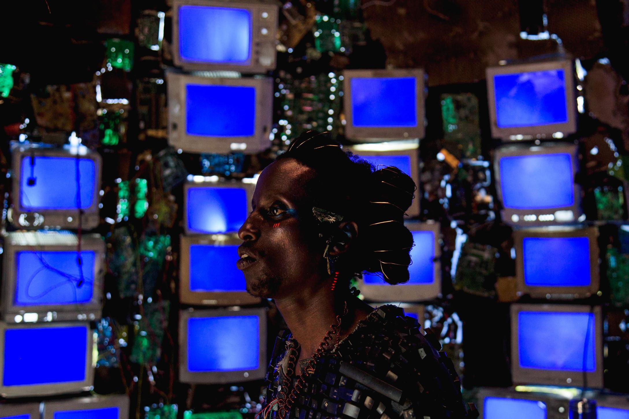 A person stands in front of a wall of old CRT monitors, each displaying a solid blue screen. They wear elaborate accessories made from electronic components, with coils in their hair and circuit boards on their clothing. The background is filled with wire