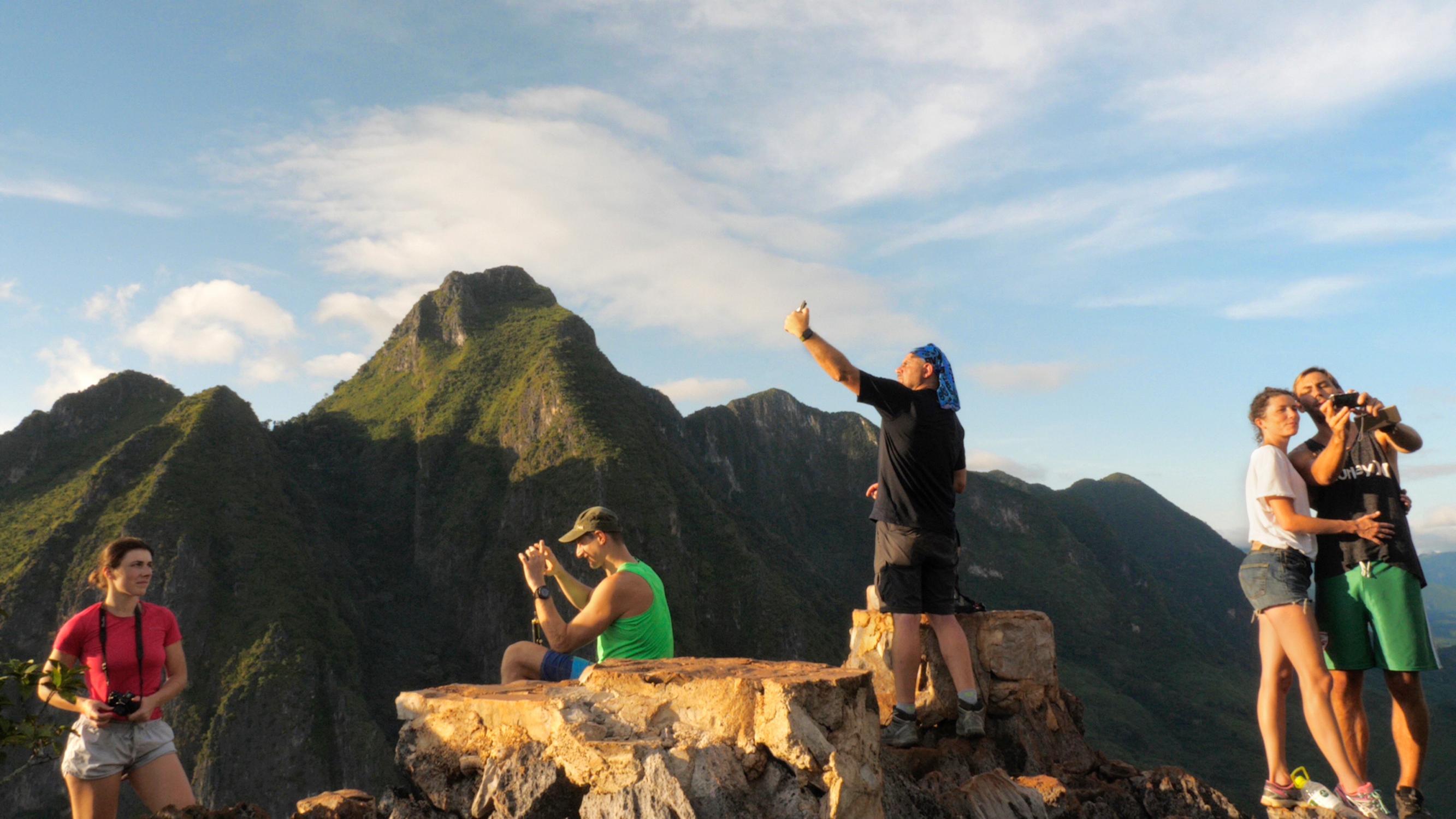 A group of people stands on a rocky mountain summit, taking photos and selfies. Behind them, lush green peaks stretch under a partly cloudy sky.
