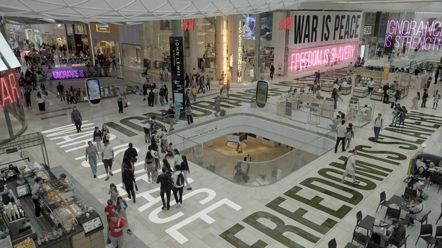 A busy shopping mall features retail stores like H&M and John Lewis, with crowds walking and dining. Bold neon slogans—“WAR IS PEACE,” “FREEDOM IS SLAVERY,” and “IGNORANCE IS STRENGTH”—are prominently displayed, evoking Orwellian themes amid the consumeri