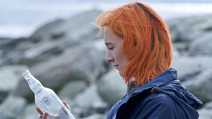 A woman with bright orange hair stares at a sand-filled glass bottle on a cold beach.