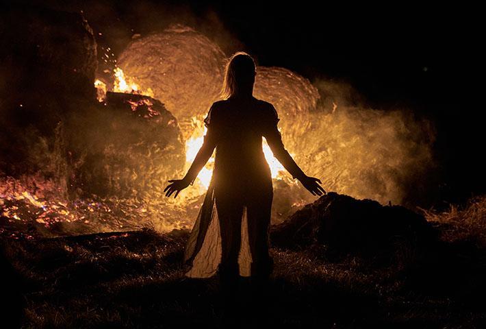 A person stands with arms slightly outstretched in front of a large fire burning hay bales at night. The flames and smoke illuminate the dark surroundings, creating a dramatic silhouette effect.