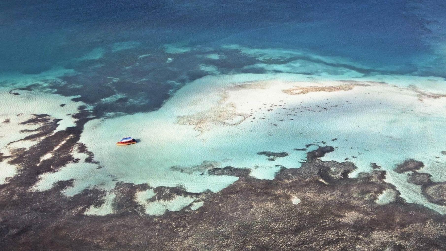An aerial view of a shallow coral reef shows a small red and blue boat anchored amid turquoise waters. Light sandy patches and darker coral formations create intricate patterns, highlighting the reef’s natural beauty and depth variations.