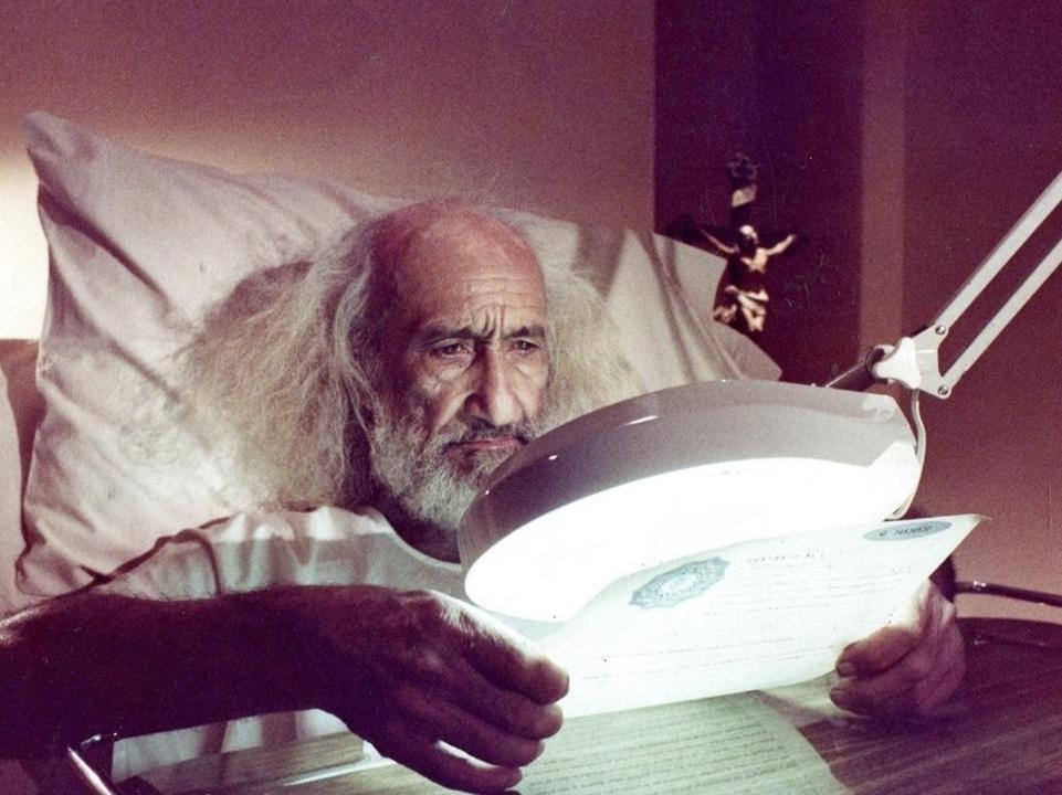 A person with long white hair sits in bed holding a document under a magnifying lamp