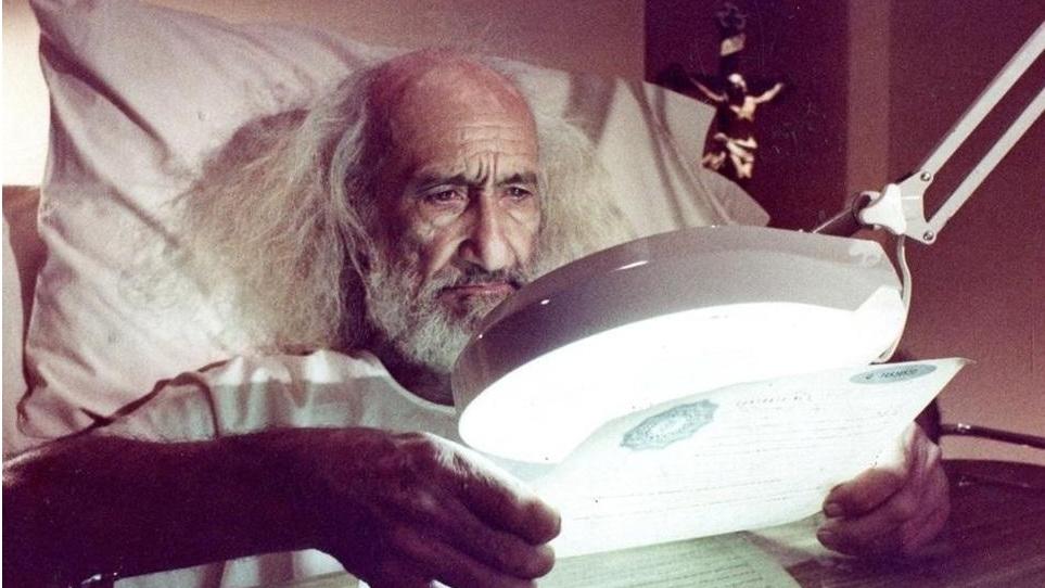 A person with long white hair sits in bed holding a document under a magnifying lamp