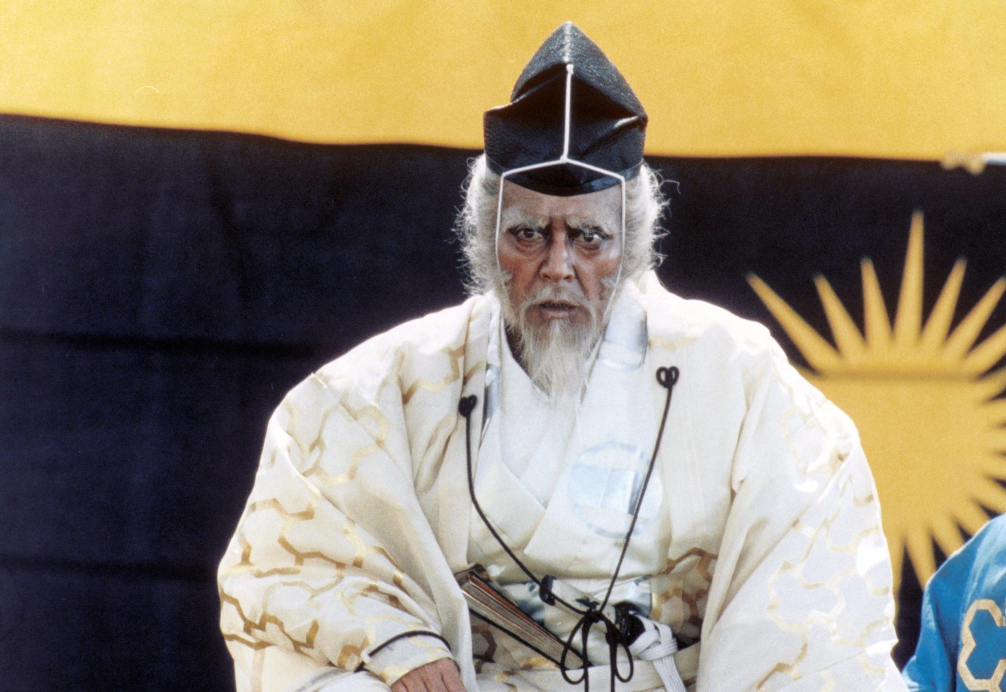 An elderly man with a white beard and intense expression wears a white robe with gold patterns and a black hat trimmed in white. He stands before a large banner featuring a yellow and black sunburst design, evoking a theatrical or historical Japanese sett