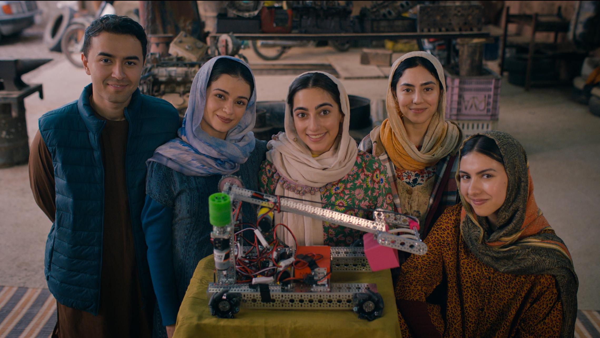 Five young people stand indoors behind a table displaying a robotic arm mounted on a wheeled base. Four women and one man wear traditional attire with headscarves and warm clothing. The background includes workshop items like an anvil, tires, and mechanic