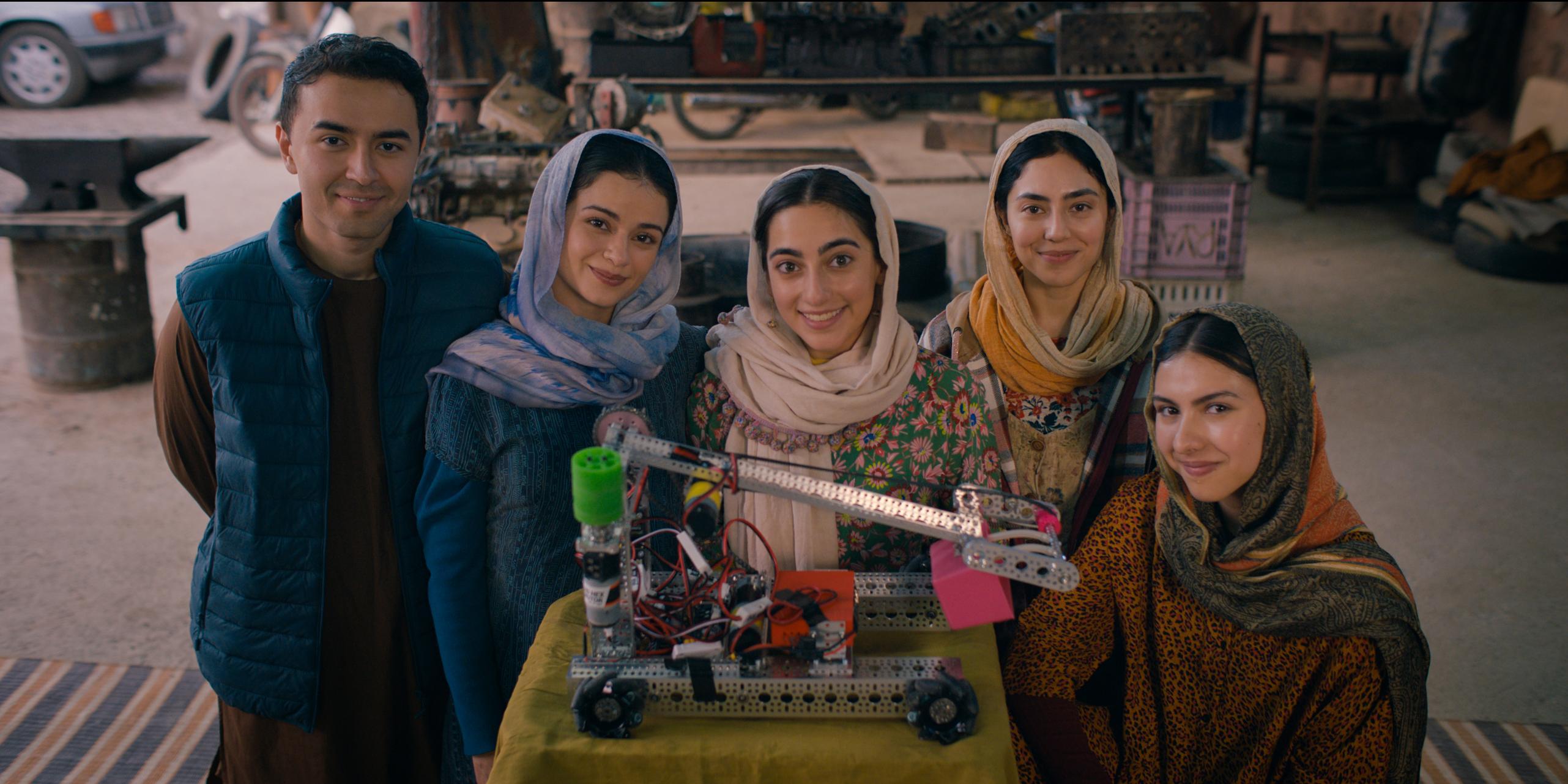 Five young people stand indoors behind a table displaying a robotic arm mounted on a wheeled base. Four women and one man wear traditional attire with headscarves and warm clothing. The background includes workshop items like an anvil, tires, and mechanic