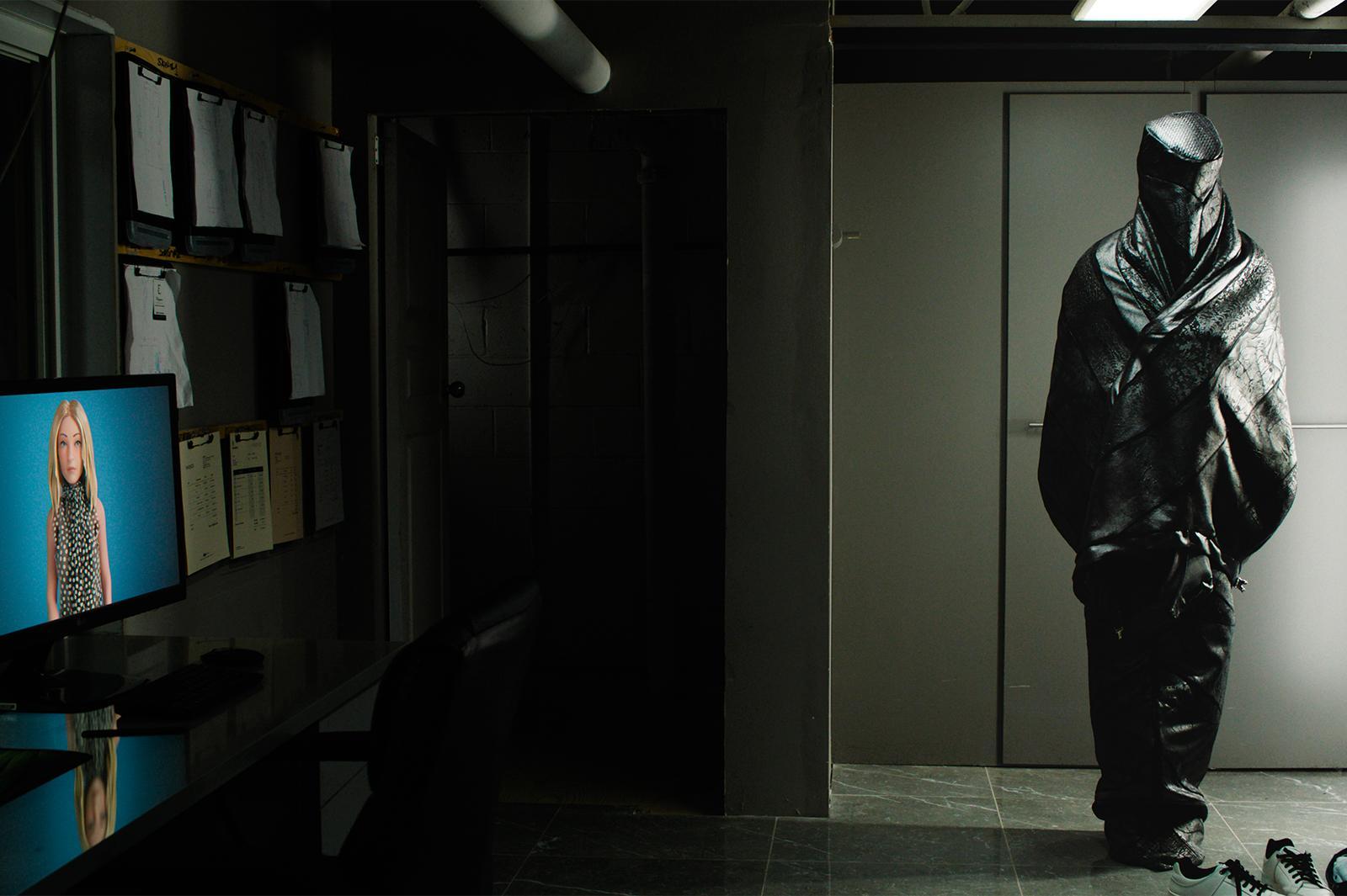 A dimly lit room shows a figure wrapped in dark, shiny material standing near a wall, while a computer monitor on a desk displays an image of a patterned garment. Clipboards are mounted on the wall to the left, and the setting appears industrial or office