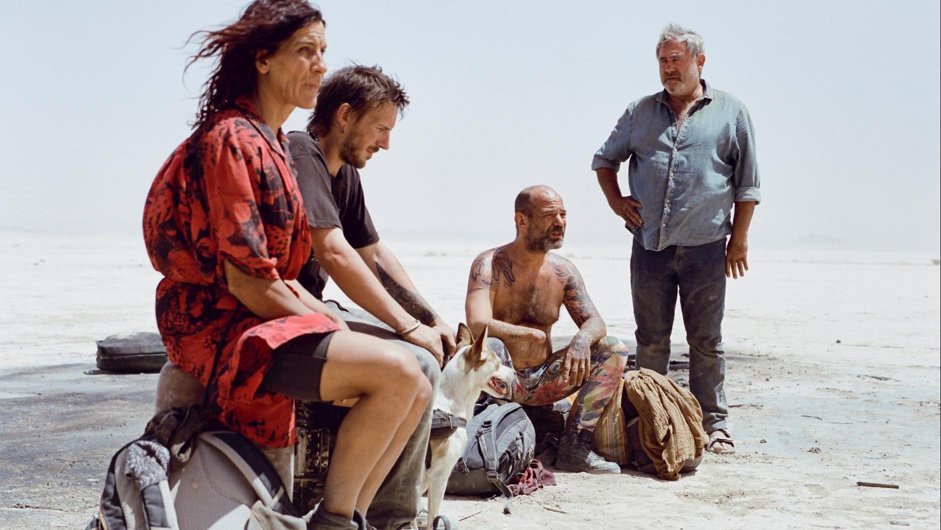 Four people and a dog gather in a desert landscape. Three sit on the ground or on bags—one in a red and black patterned outfit, one in a black shirt, and one shirtless with tattoos and colorful pants. A fourth person stands nearby in a blue shirt and jeans. Backpacks and gear surround them, and the clear sky emphasizes the stark, sandy terrain.