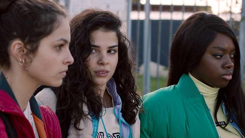 Three people stand outdoors near a metal fence, dressed in jackets and sweaters. One wears a red jacket, another has curly hair and a light hoodie, and the third wears a green jacket over a yellow turtleneck. Their serious expressions suggest a moment of 