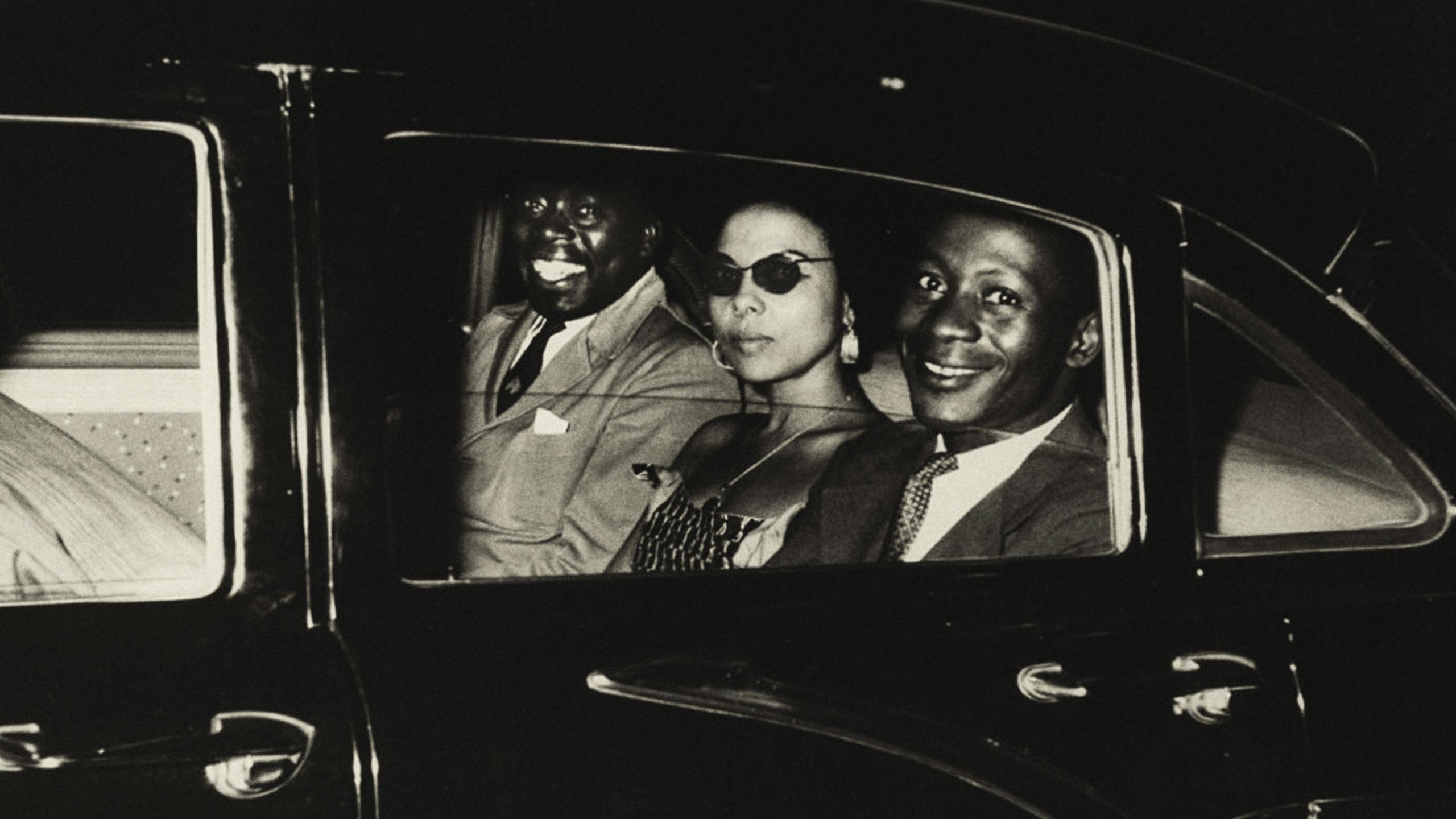 A black-and-white photo shows three formally dressed individuals seated in the back of a car at night. Two men wear suits and ties, and a woman in a strapless dress and sunglasses sits between them.