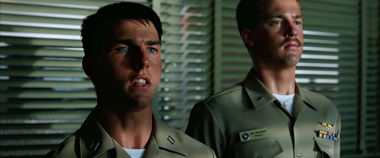 Two individuals wearing military uniforms stand indoors in front of horizontal window blinds. One uniform displays a name tag, insignia, and colorful ribbons.
