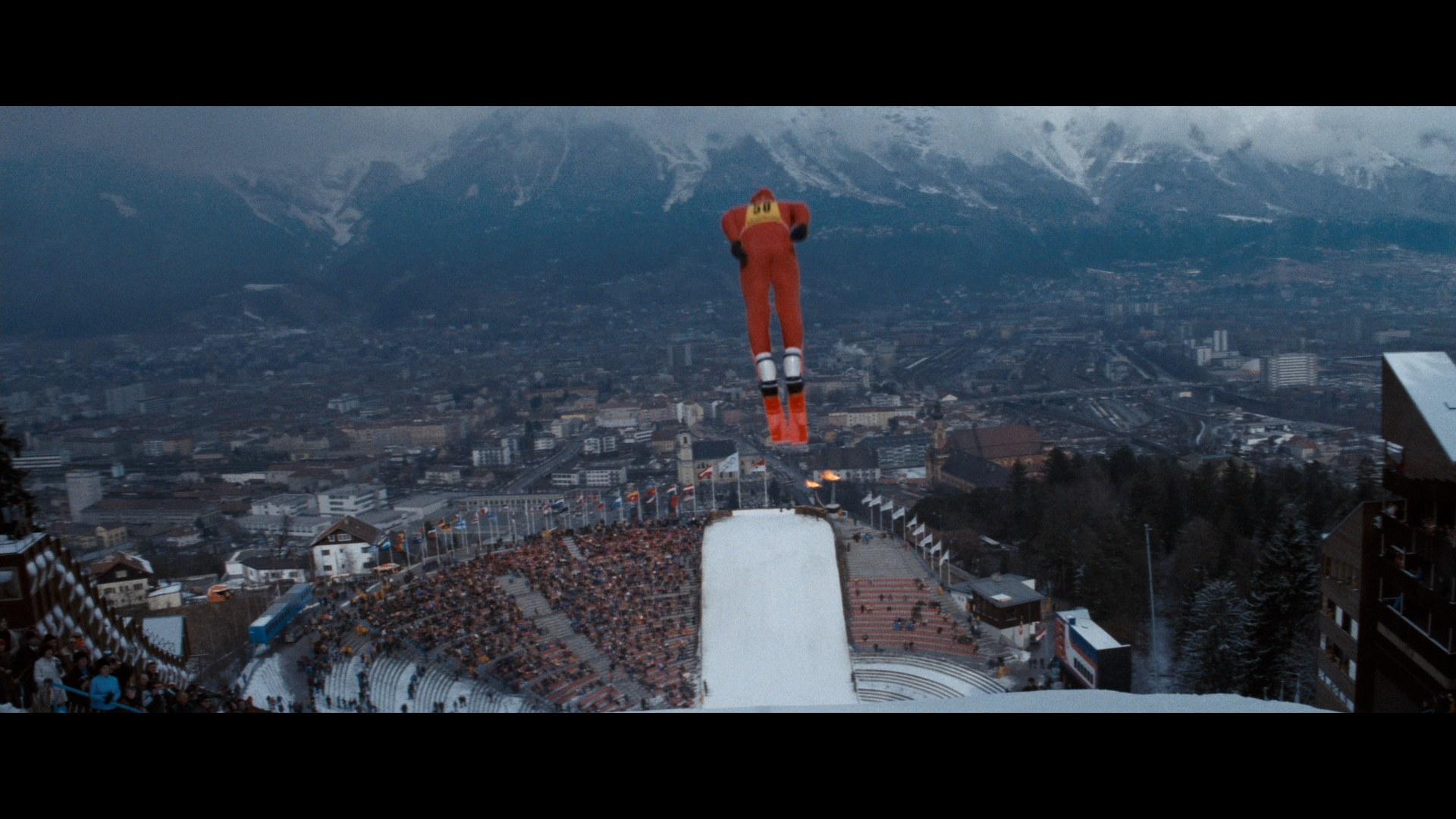 A ski jumper in a blue suit is airborne above a snowy ramp, with a large crowd in a stadium below and a cityscape surrounded by mountains in the background. The scene captures the moment of flight during a ski jumping event.