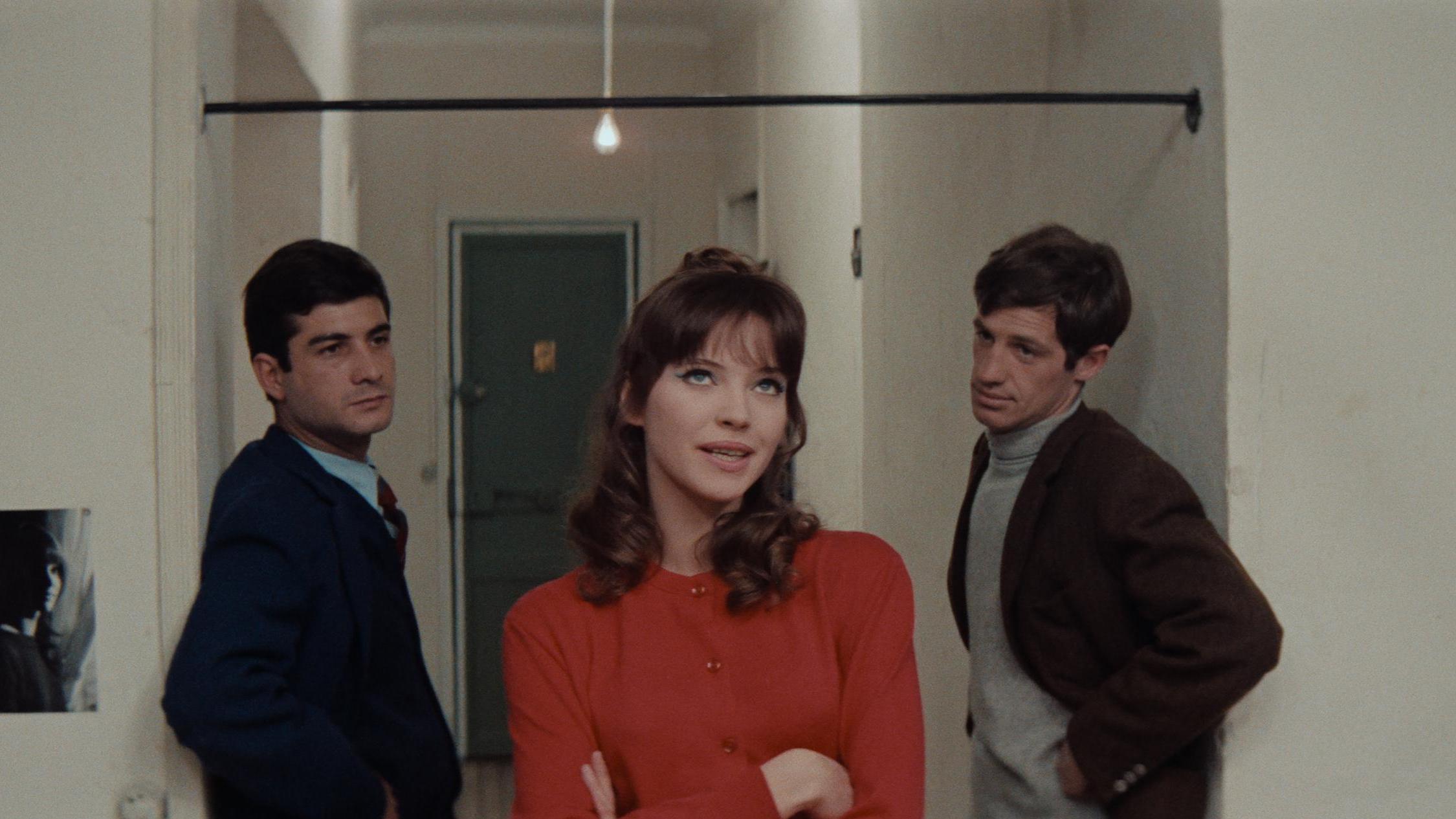 Three people stand in a hallway with white walls and a green door, engaged in a tense conversation. The central figure in red gestures while the others listen.