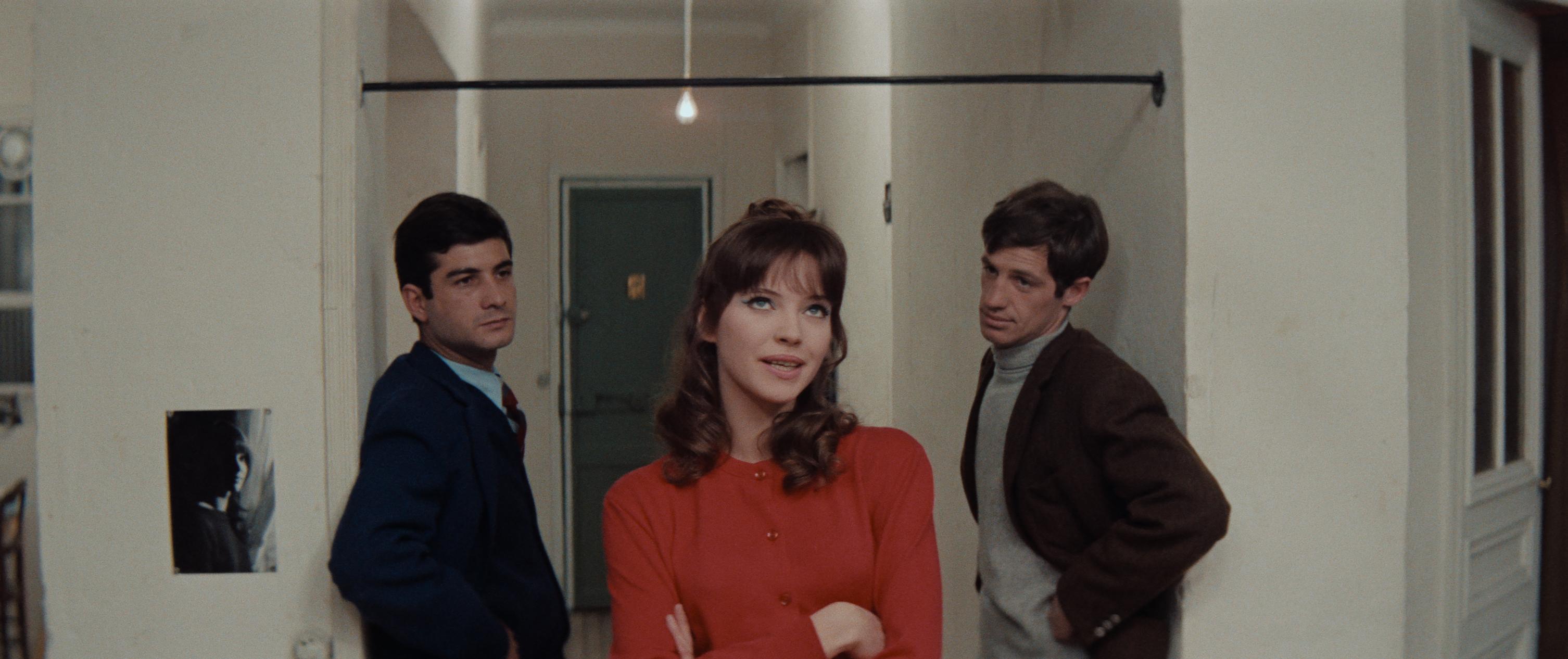 Three people stand in a hallway with white walls and a green door, engaged in a tense conversation. The central figure in red gestures while the others listen.