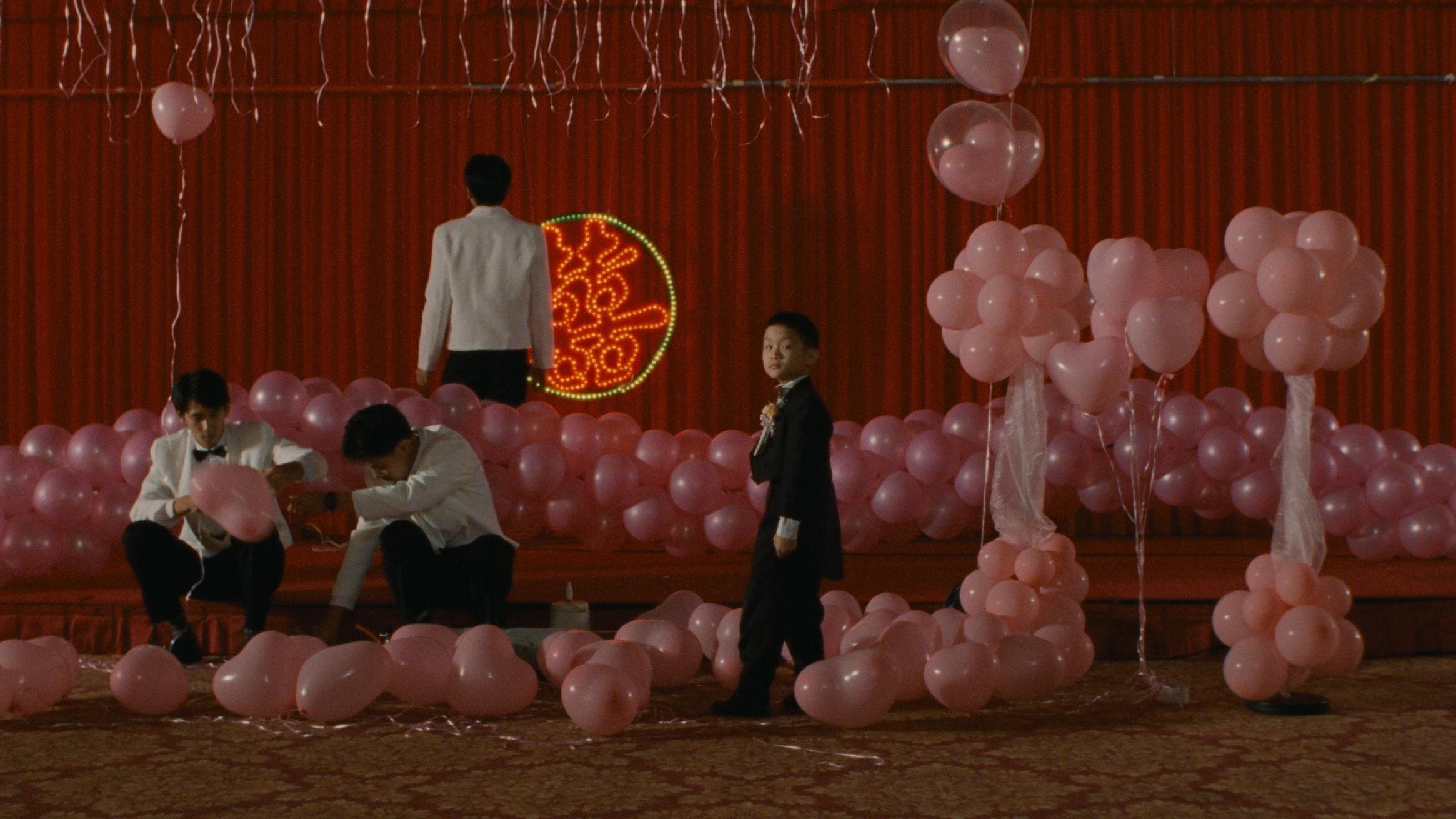 A decorated event space with pink balloons arranged in clusters and scattered on the floor, featuring people in formal attire. A red backdrop with a circular neon design is visible behind the stage area.