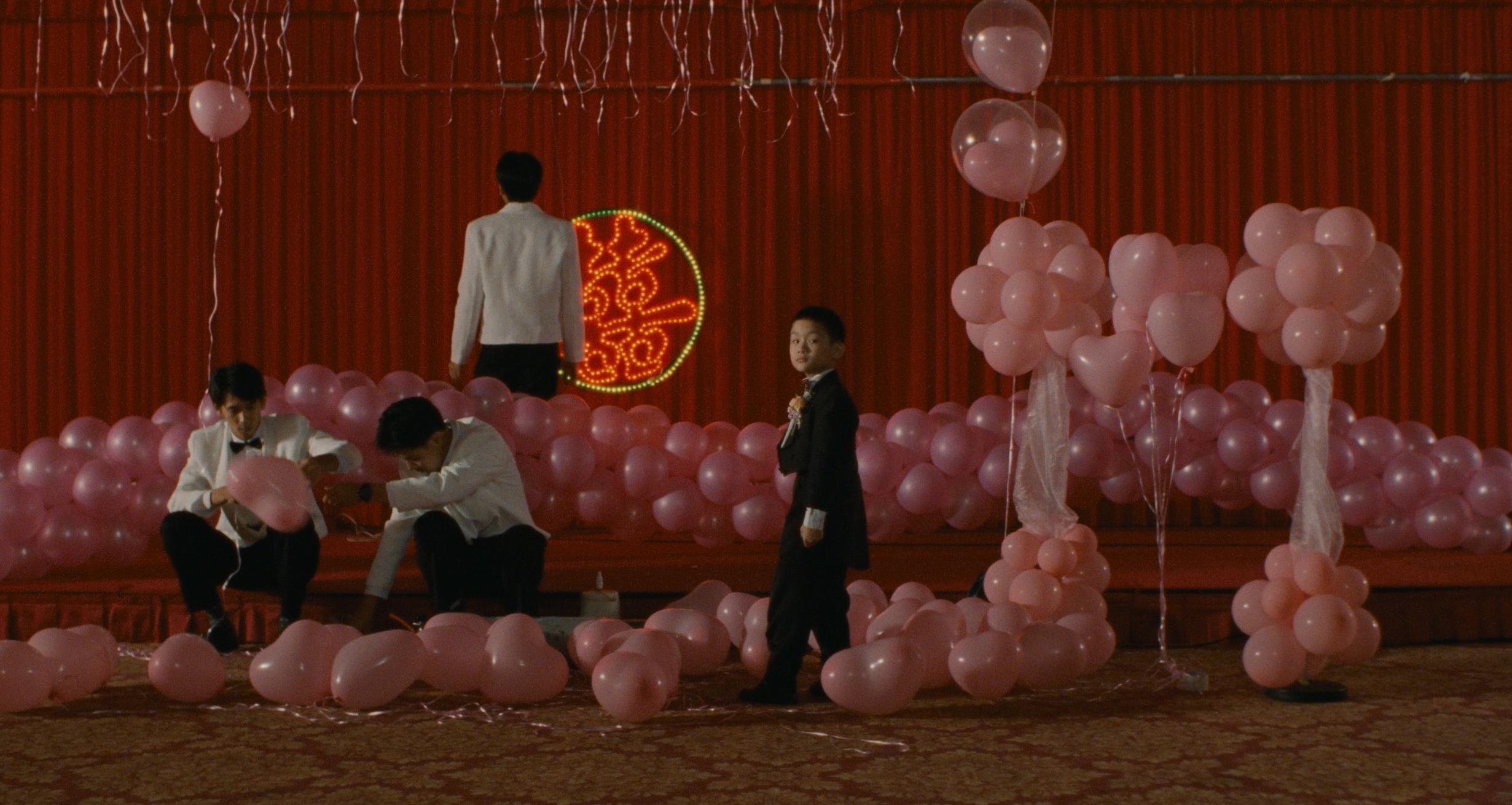 A decorated event space with pink balloons arranged in clusters and scattered on the floor, featuring people in formal attire. A red backdrop with a circular neon design is visible behind the stage area.