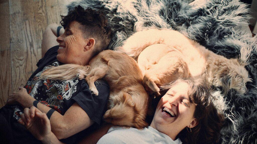 A close-up photo of a couple laughing while laying in the sunshine cuddling with two fluffy pets.
