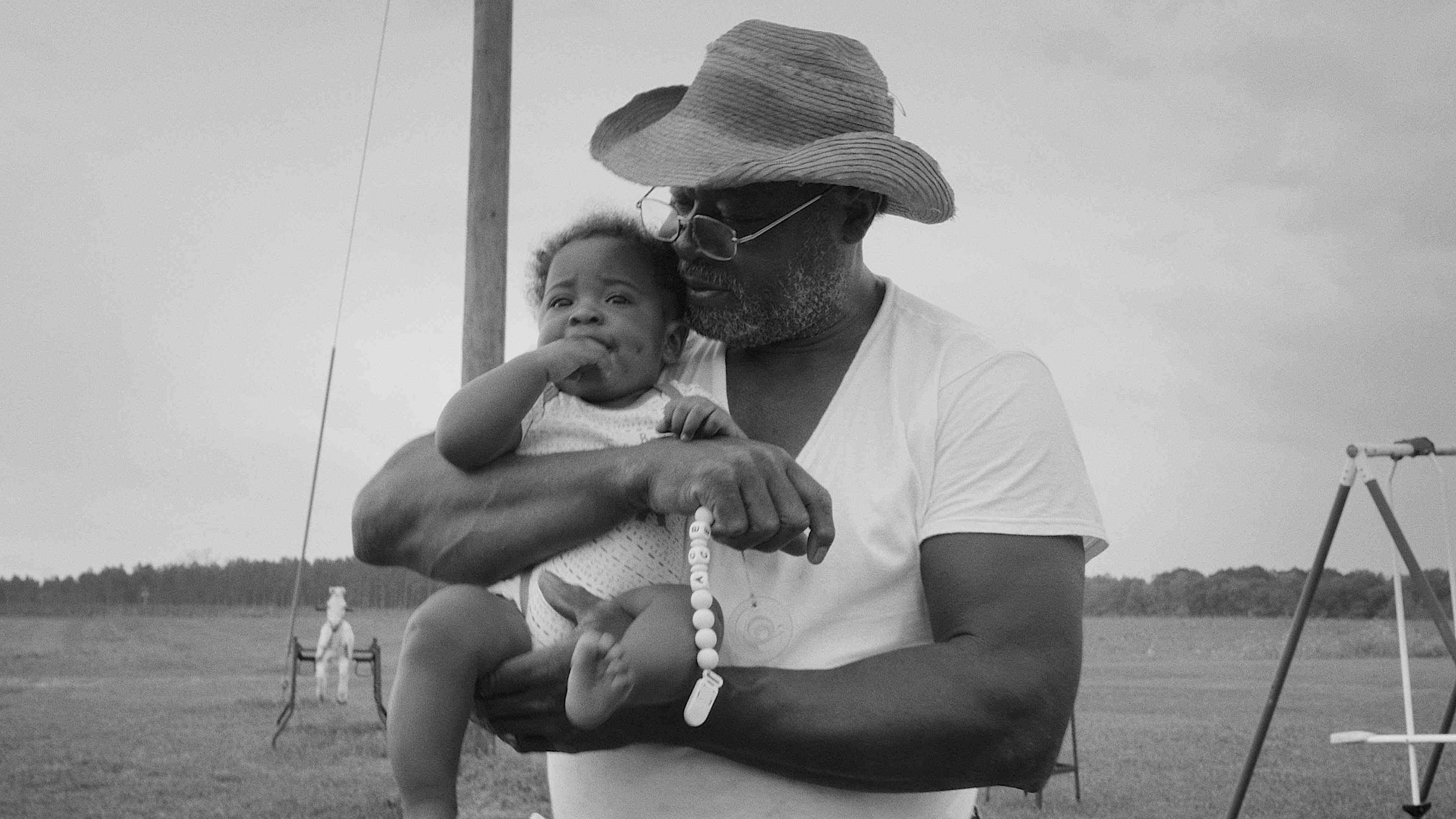 A monochrome image of an older man wearing a cowboy hat and white t-shirt holding a chubby baby.