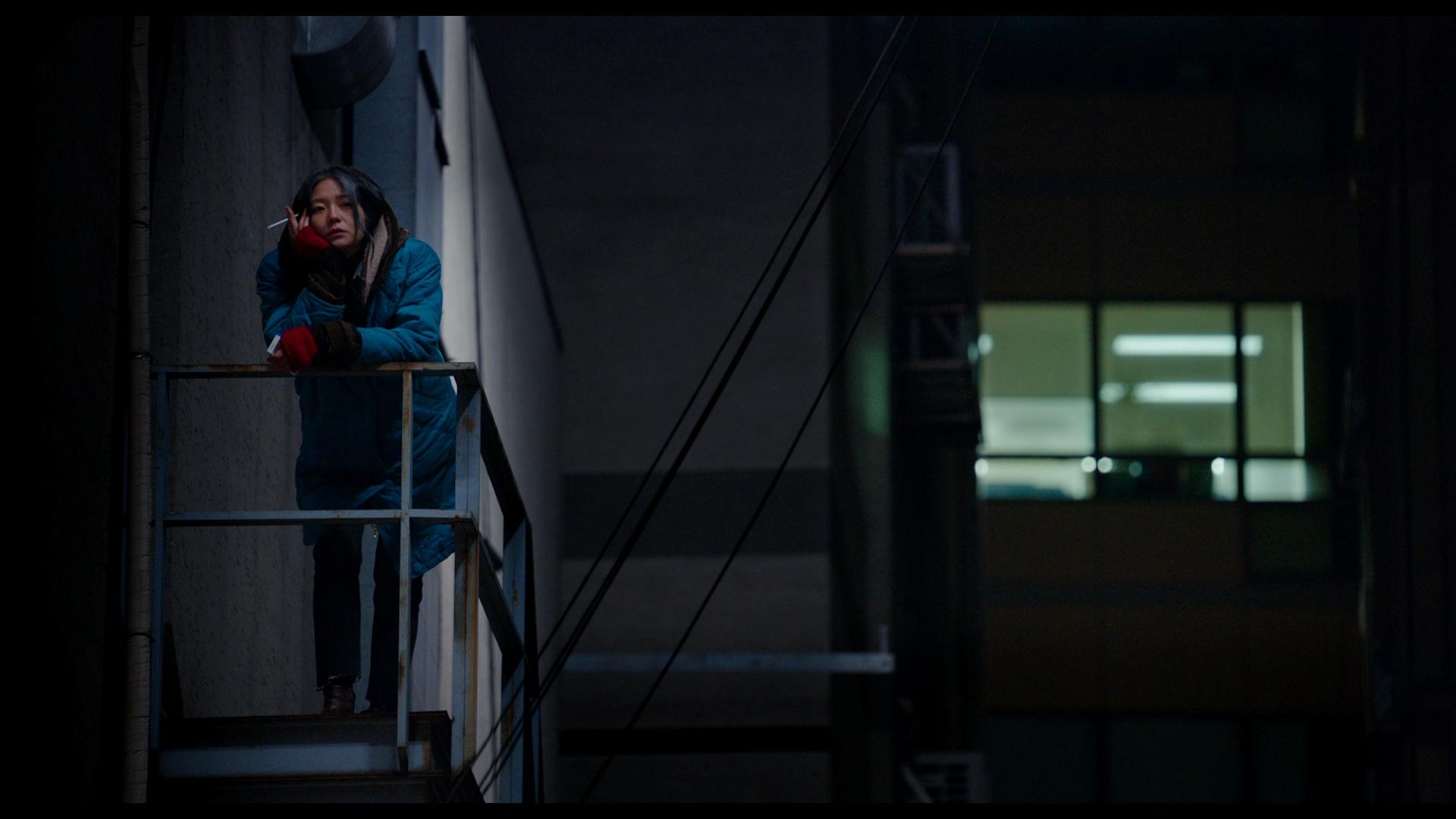 A young woman standing on a fire escape at night smoking a cigarette.