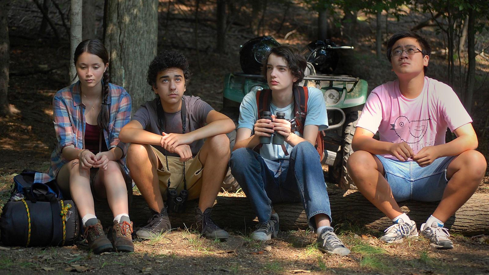 Four teenagers are sitting on a log in the woods. One of them is holding a pair of binoculars and one is looking up at the tree tops.