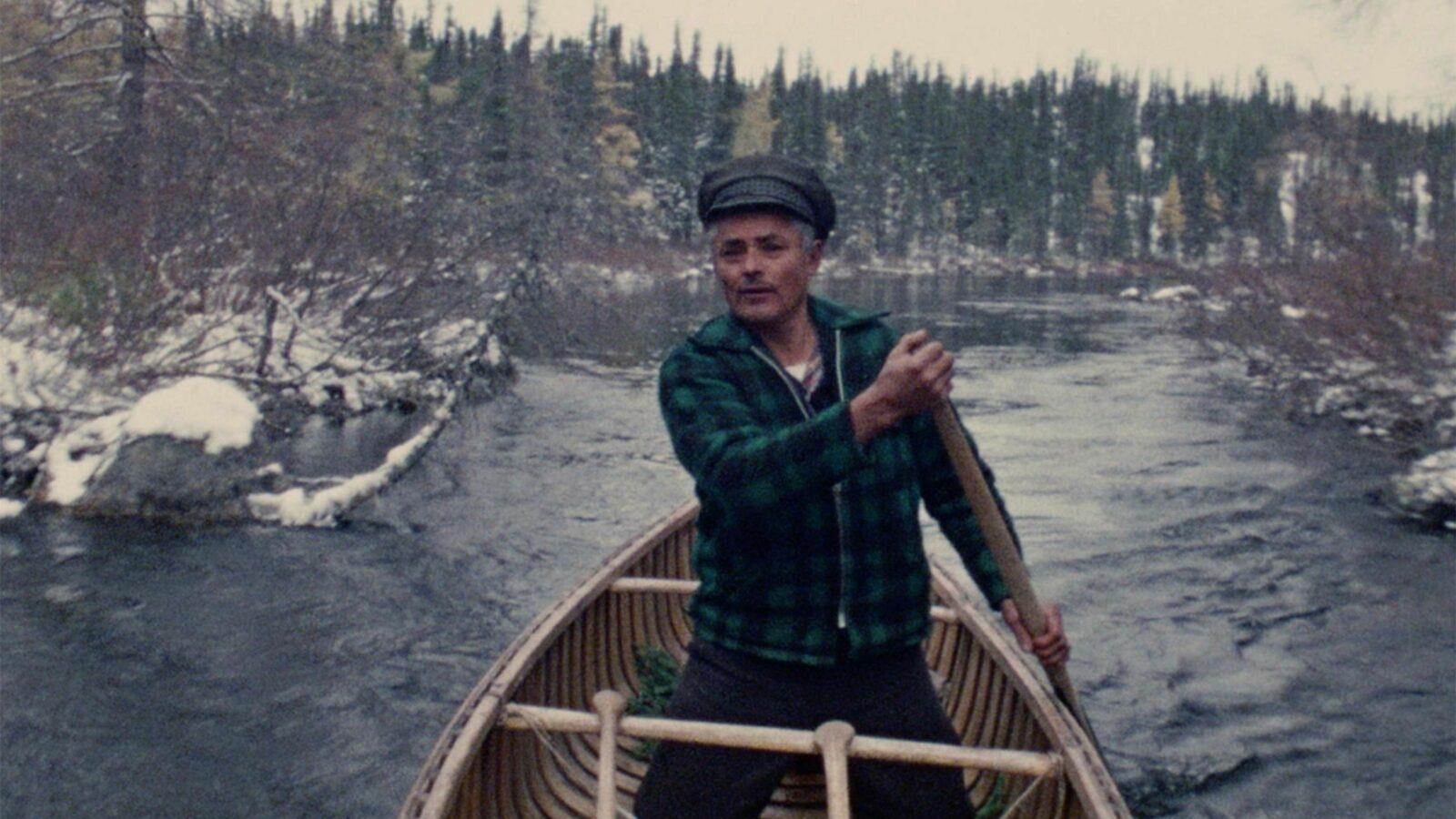 A man wearing a green plaid shirt and a hat rows a boat down a river surrounded by a snowy forest.
