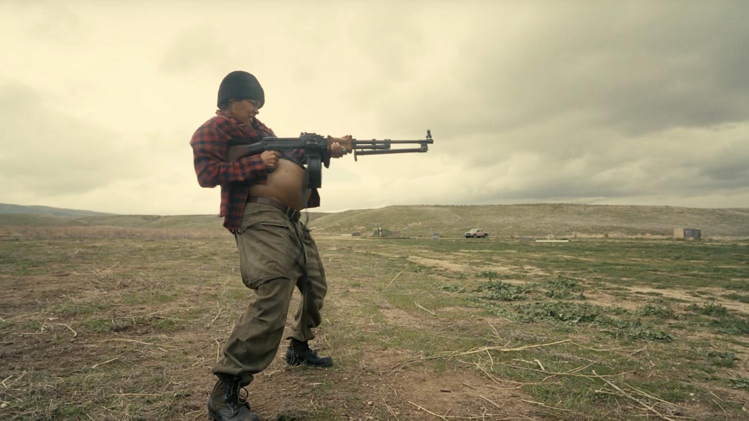 A woman wearing a knit hat and plaid flannel shirt with an exposed pregnant belly shooting a machine gun in an empty field.