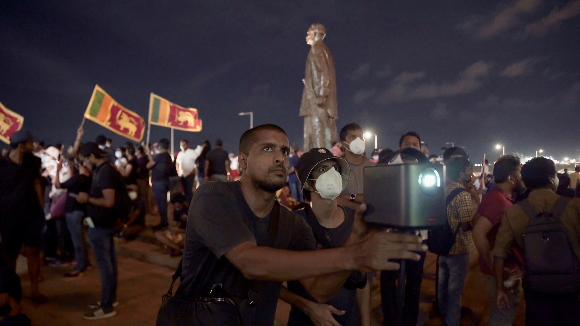 A man and a boy hold a camera at a protest. There is a crowd of people holding flags and a tall statue of a person in the background. The boy is wearing a white mask over his mouth. 