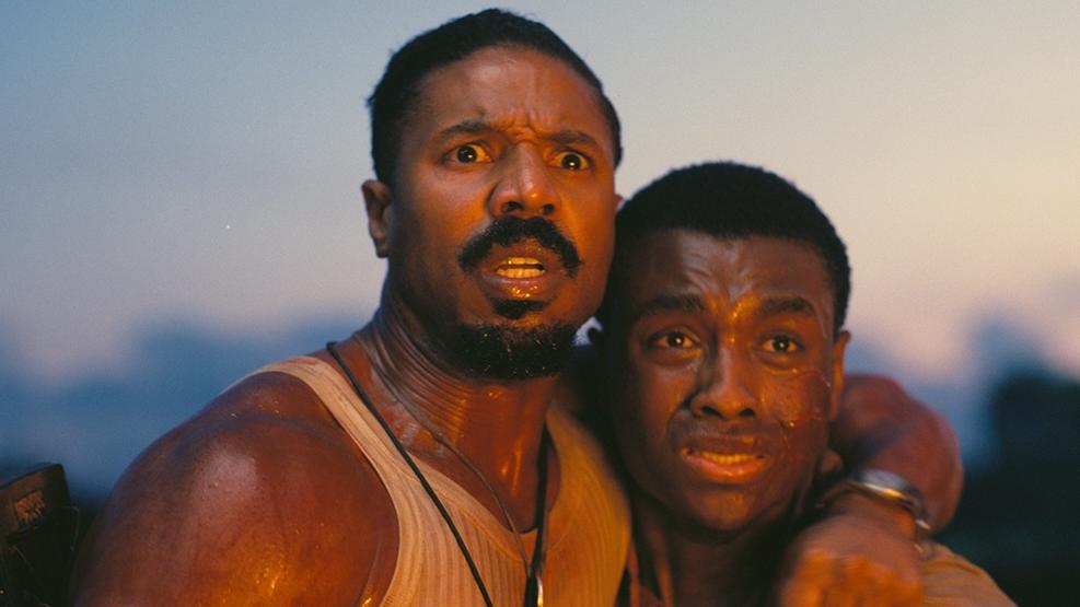 Two men clutch each other with fearful looks on their faces. They appear to be illuminated by fire light.