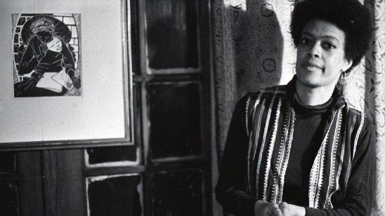 A black-and-white photo of a woman wearing a striped vest. She is leaning against the wall on the right side of the image. At the left, there is a painting on the wall.