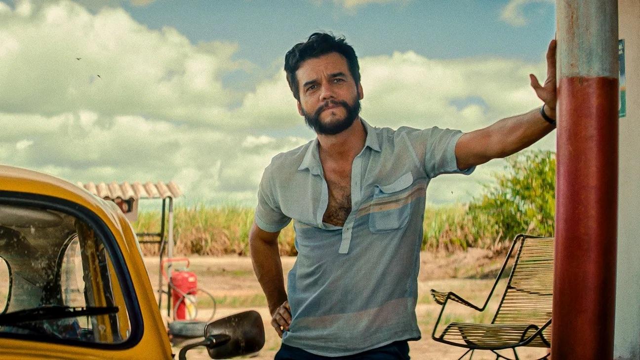 A man with a beard wearing a blue shirt leaning with one hand against a pole at what looks like a gas station. There is a yellow car to his right and it appears to be an arrid climate.