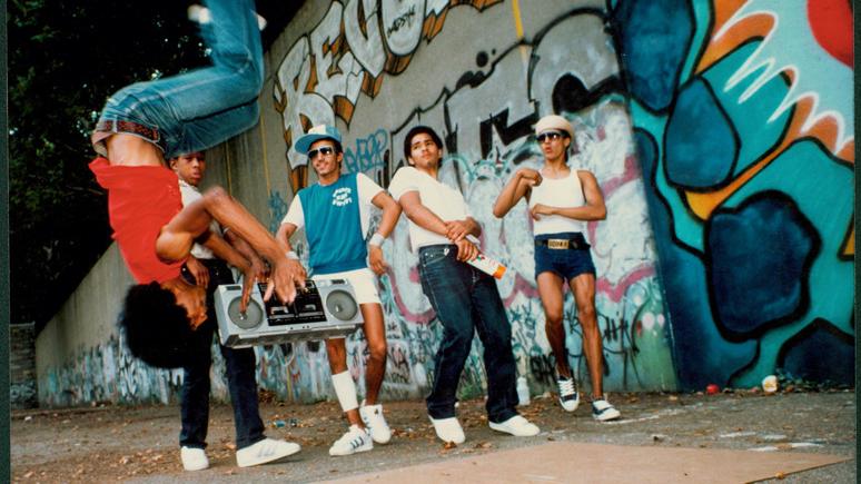 A group of people dancing in front of a wall covered in bold graffiti. One of the people in the air doing a backflip and another is holding a large boom box.