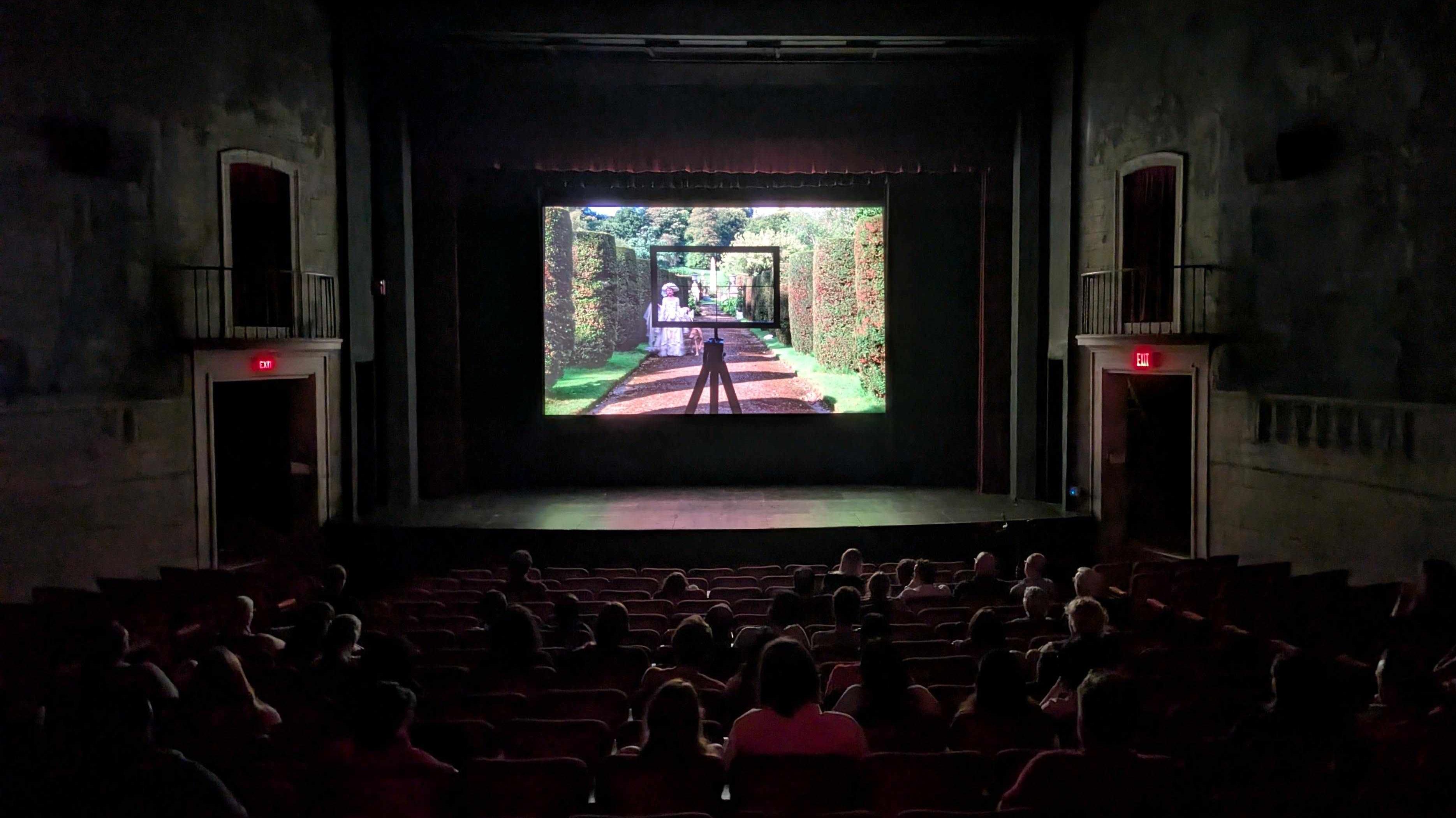 A group of people inside a darkened movie theatre with an image of a woman in a garden wearing a frilly white dress framed by a black drawing instrument.