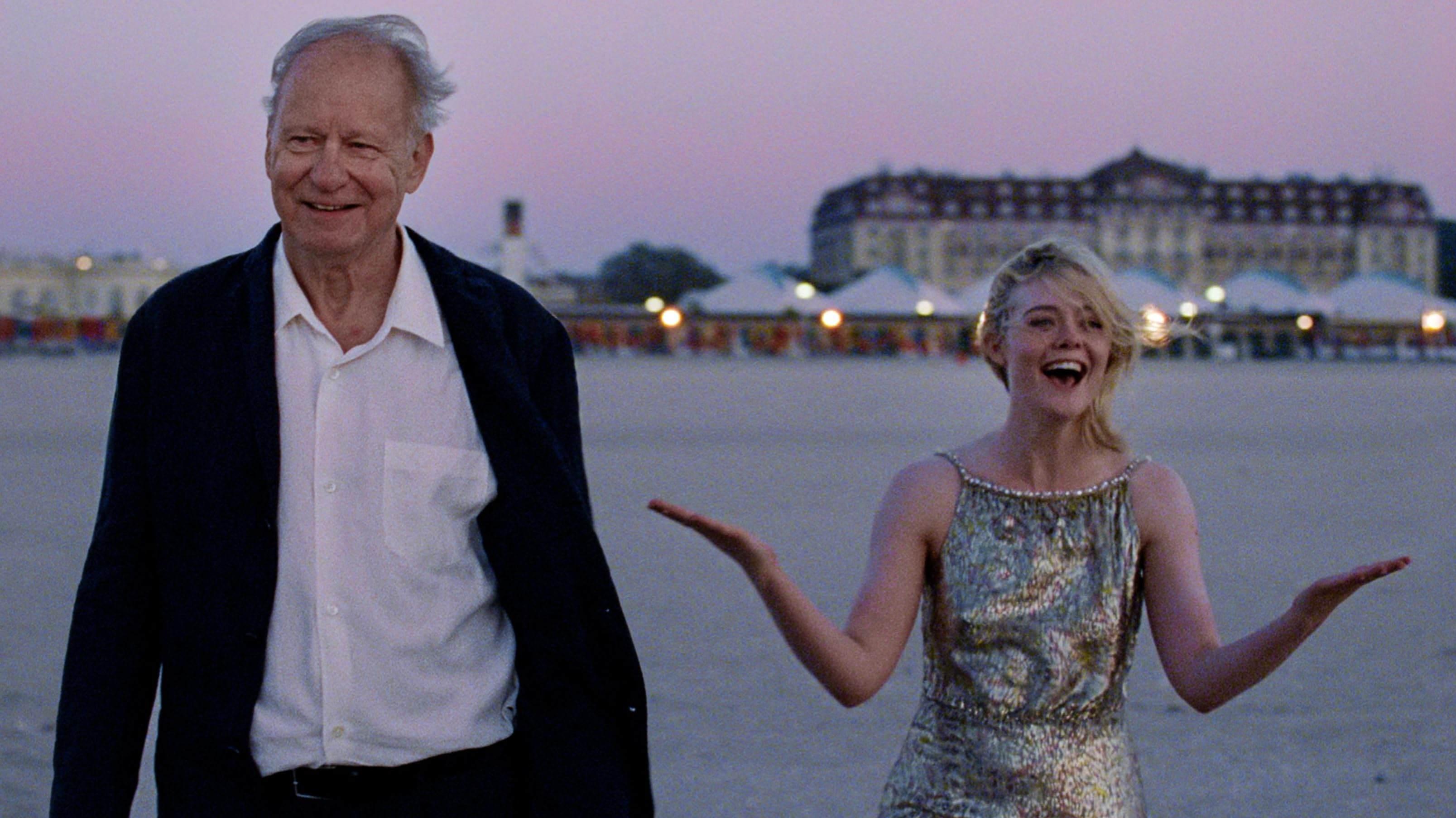 An older man wearing a suit walks on a beach at dusk beside a young blonde woman wearing a sparkly silver dress who is gesturing joyously.