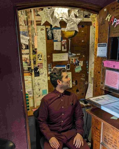 A man in a maroon shirt sits inside a small office with decorations all over the walls.