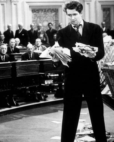 A forlorn looking man standing in the center of a courtroom holding a disorganized stack of papers in his hand.