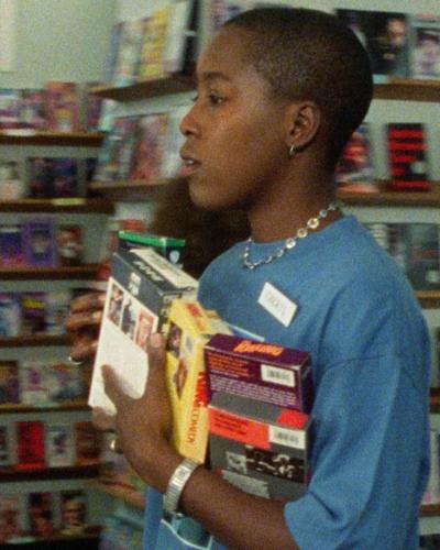 A grainy image of woman with a blue shirt carrying an armful of VHS tapes inside a video store.