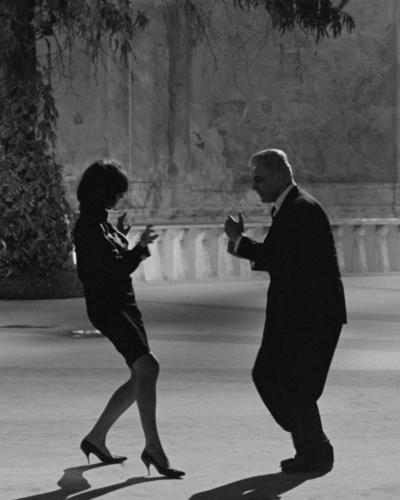 Two people dance energetically in an open courtyard, one in a dress and heels, the other in a suit. A third person sits at a table under a canopy, watching the scene unfold.